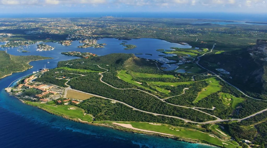 Aerial view of Old Quarry Golf Course in Curaçao, showcasing the lush green fairways, surrounding hills, water bodies, and nearby marina under a partly cloudy sky including the Sandals Royal resort.